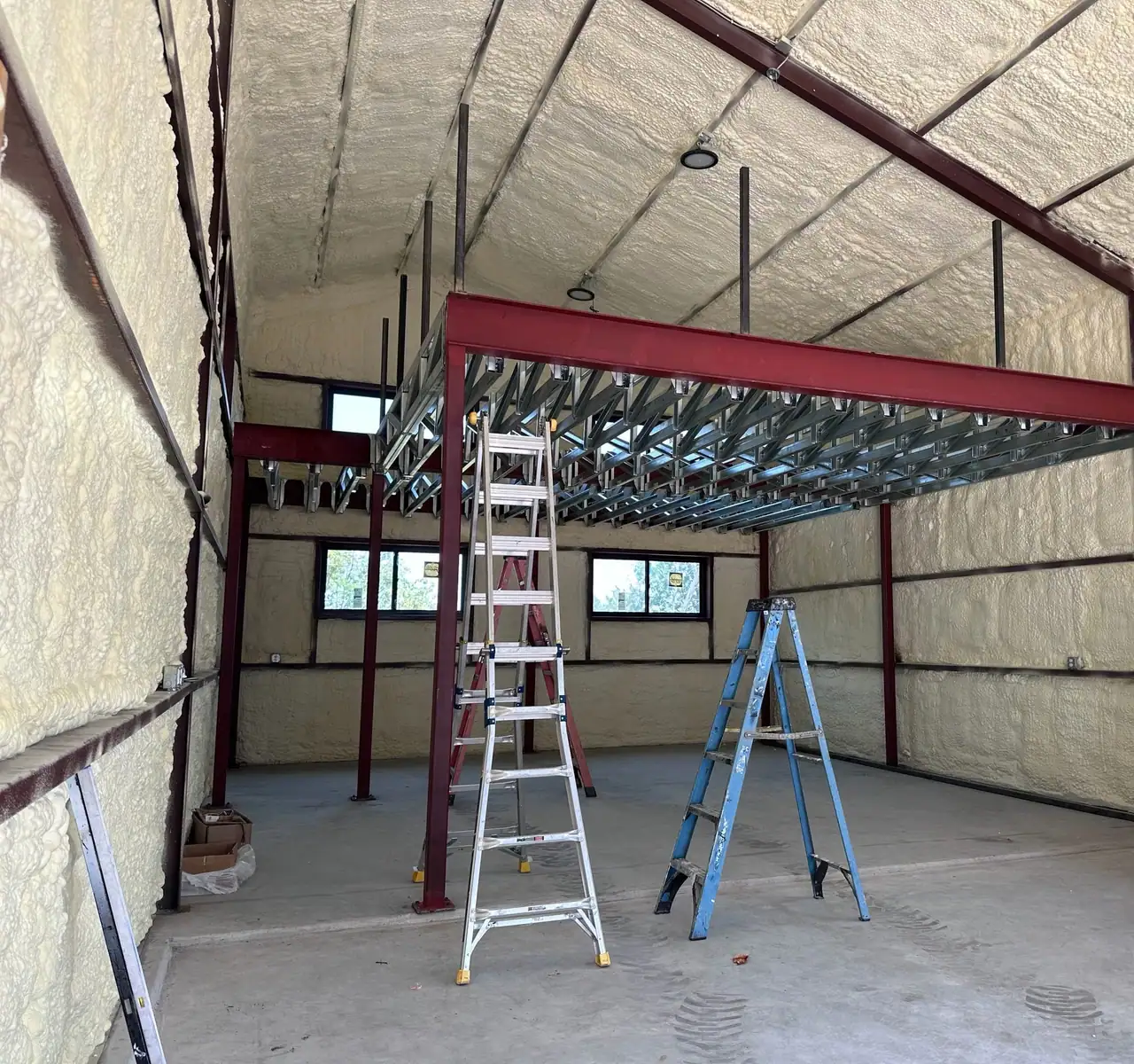 Steel building interior with spray foam on walls and roof, a red steel mezzanine frame with metal joists, and ladders