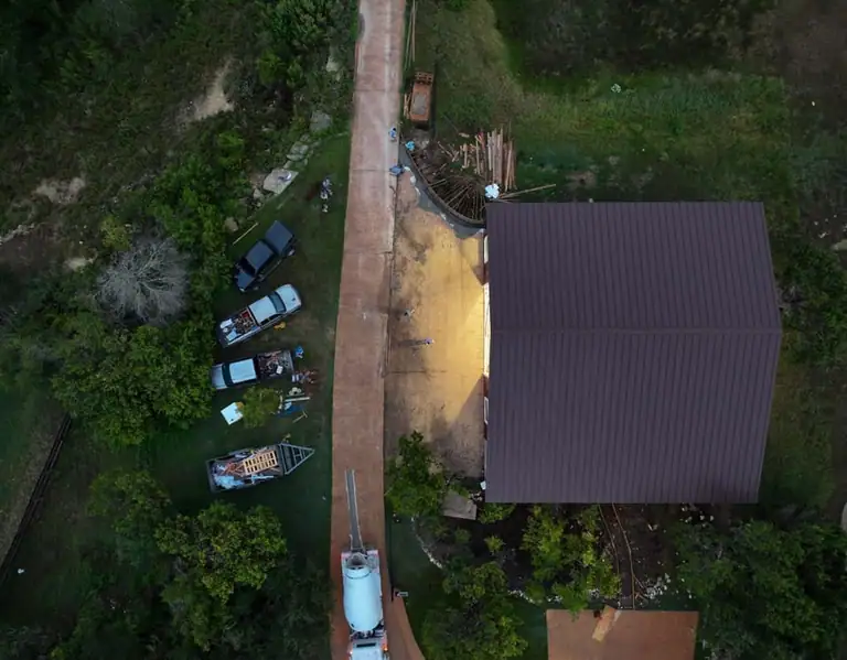 Straight-down drone view of the red shop's brown metal roof, rebar grid and a mixer truck on a stained concrete drive