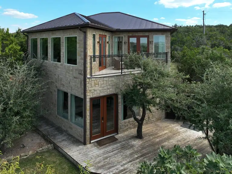 Two-story stone guest house with metal roof, iron-railed balcony, wood French doors and a wood deck among trees
