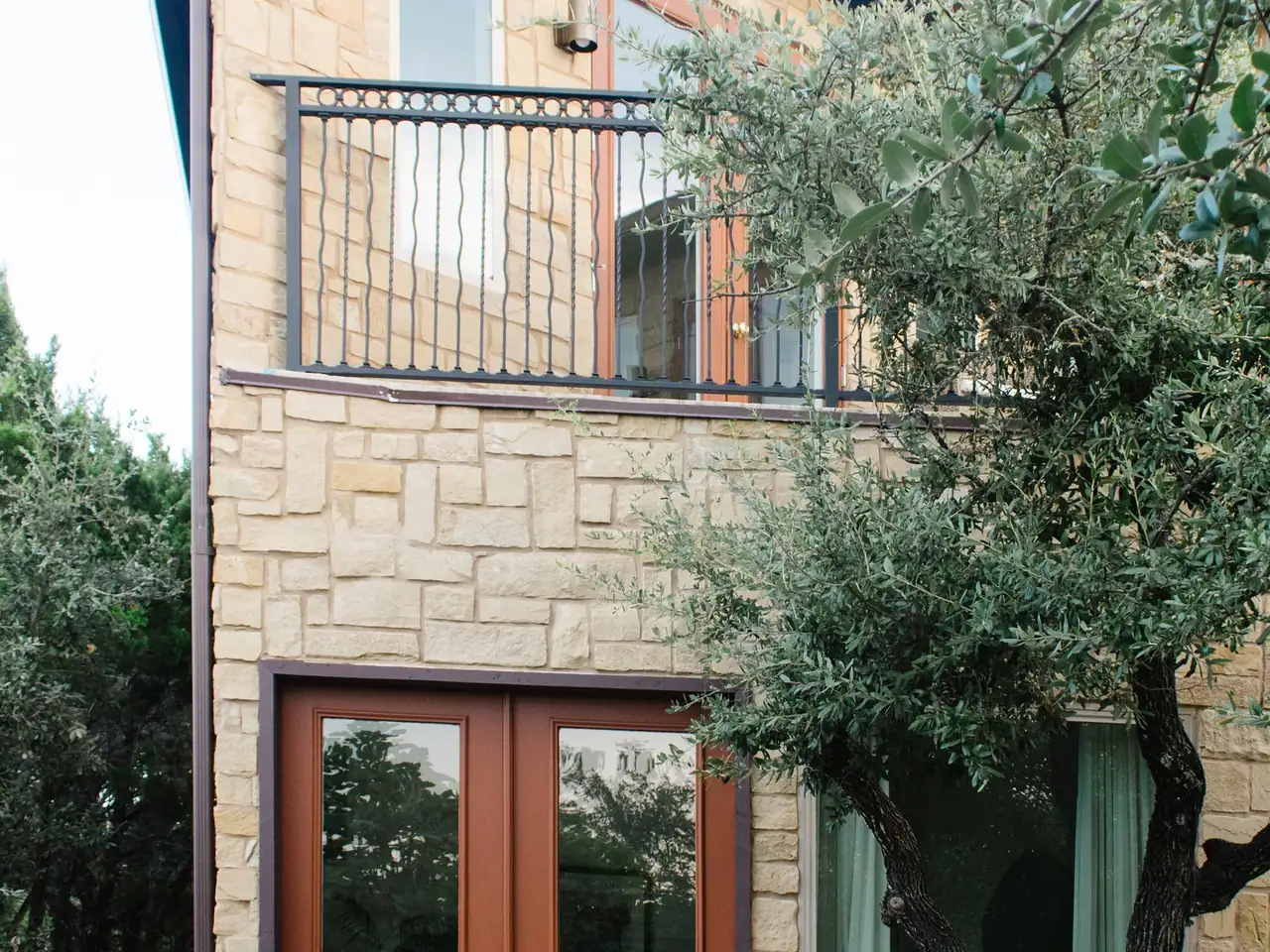 Stone guest house wall with an iron balcony, wood-tone French doors and an olive tree in front
