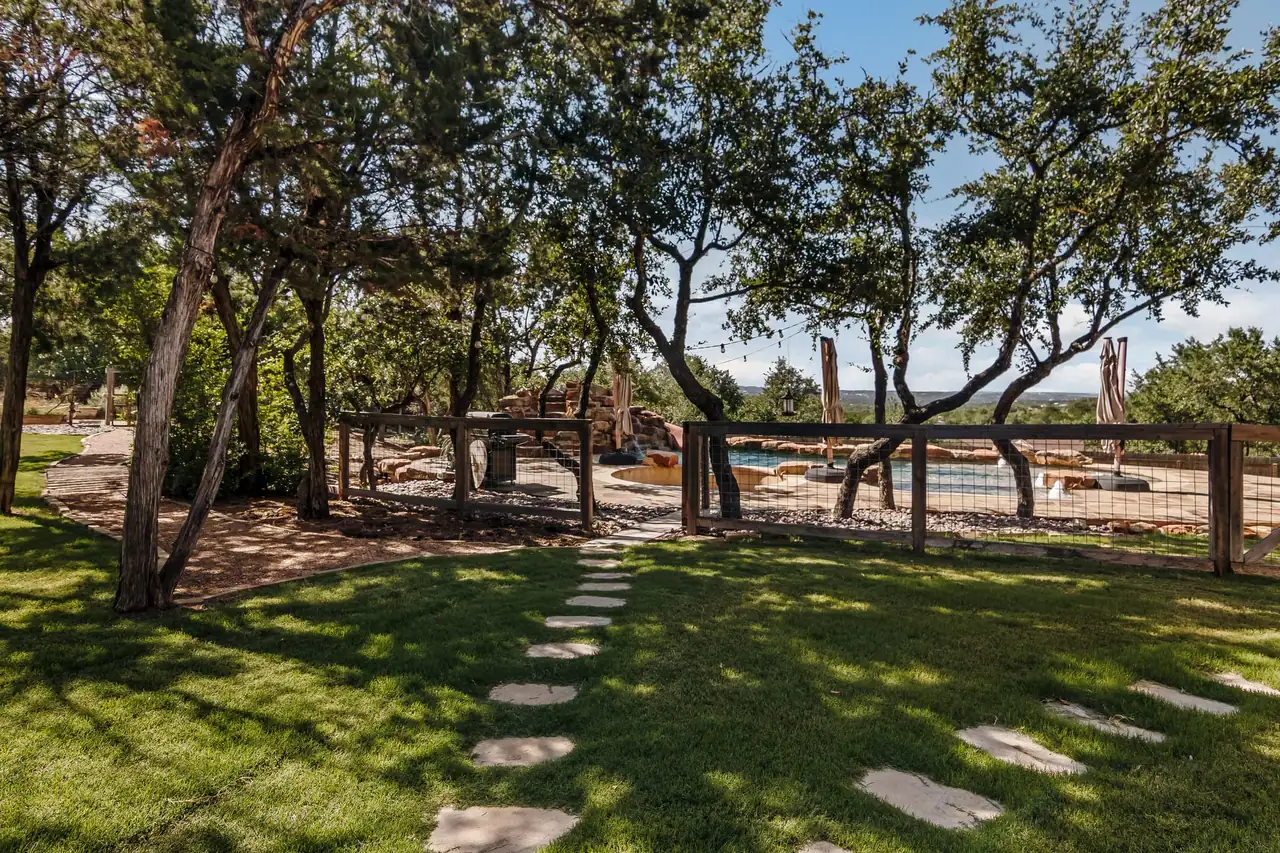 Lawn with flagstone stepping stones leading through oaks to a wood-and-wire pool fence, with a boulder-edged pool beyond