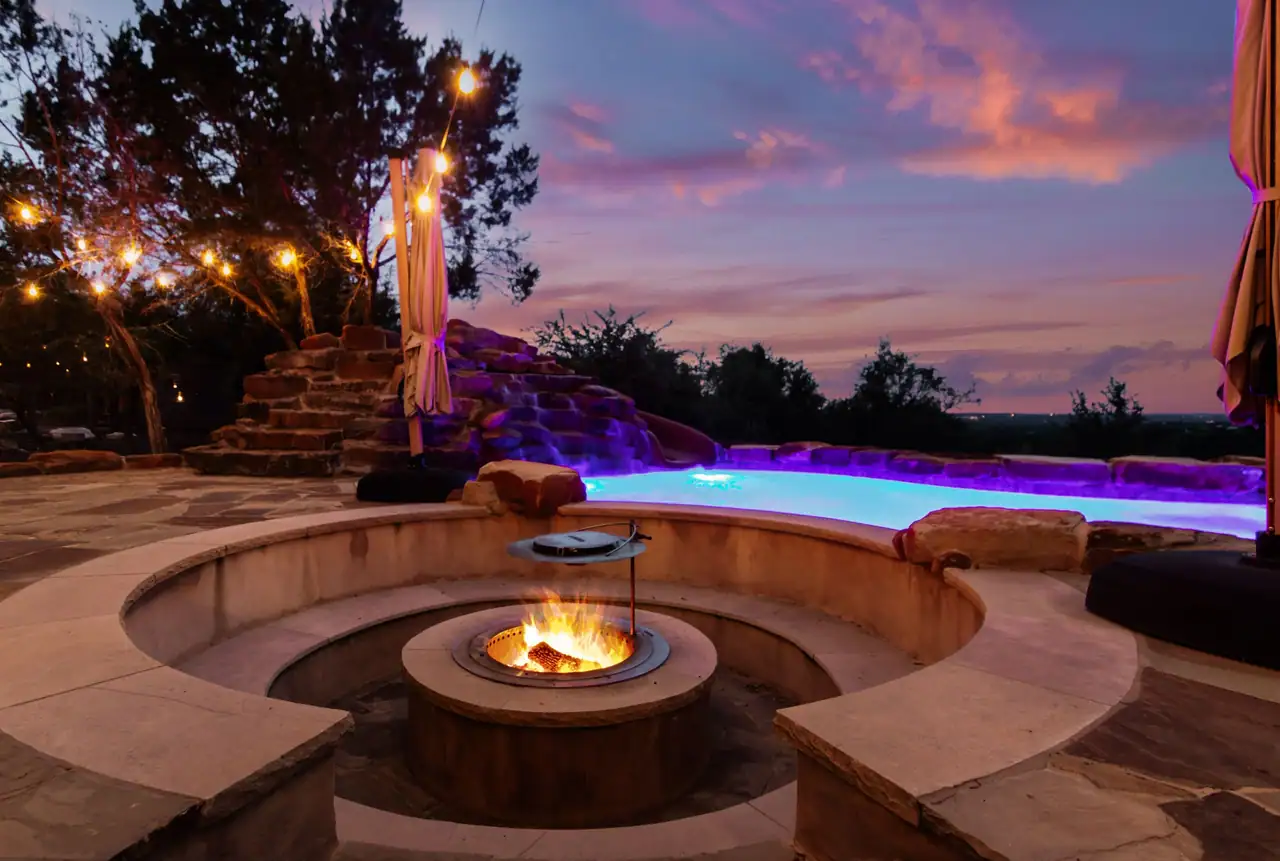 Sunken round fire pit with a curved stone bench and a lit fire beside a glowing pool and rock waterfall at dusk