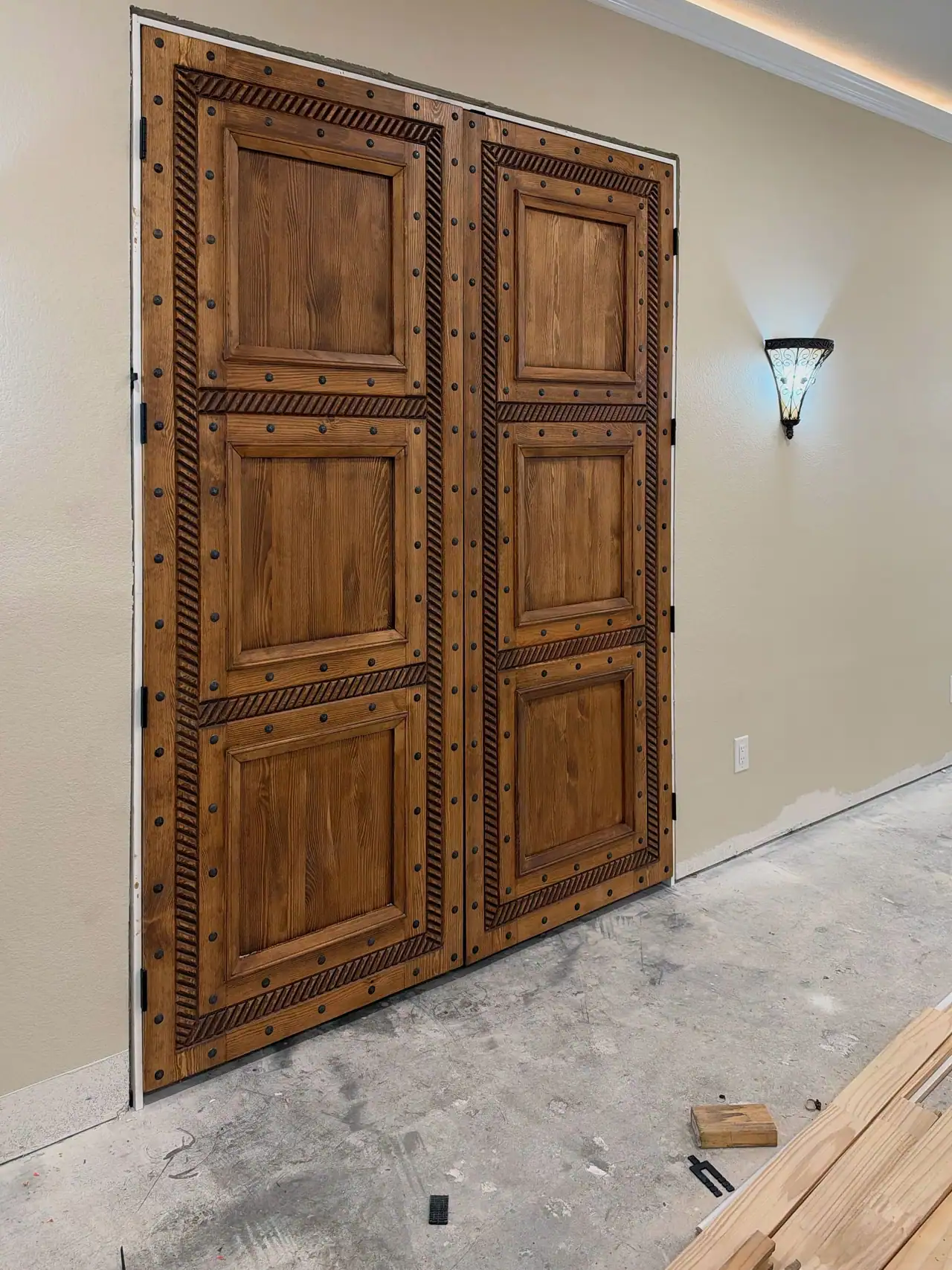 Rustic stained wood double doors with carved rope trim and round metal studs, lit glass wall sconce, concrete floor