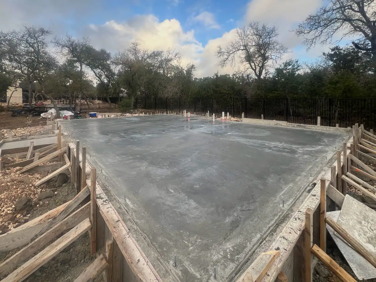 Freshly poured concrete slab in wood forms with stakes and braces, iron fence and oak trees under cloudy morning sky