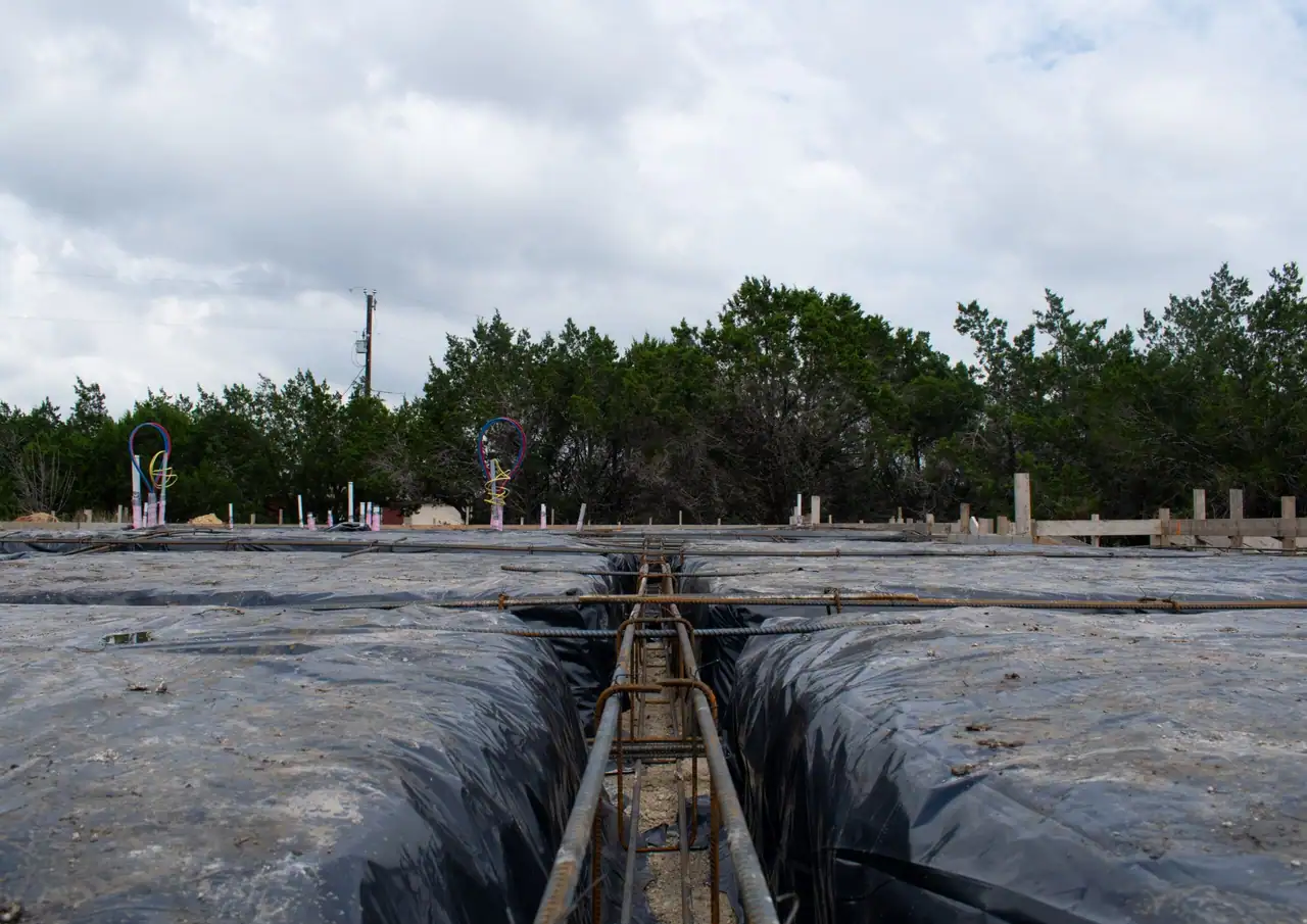 Rebar cage running down a beam trench lined with black vapor barrier, plumbing stub-outs and cedar trees beyond