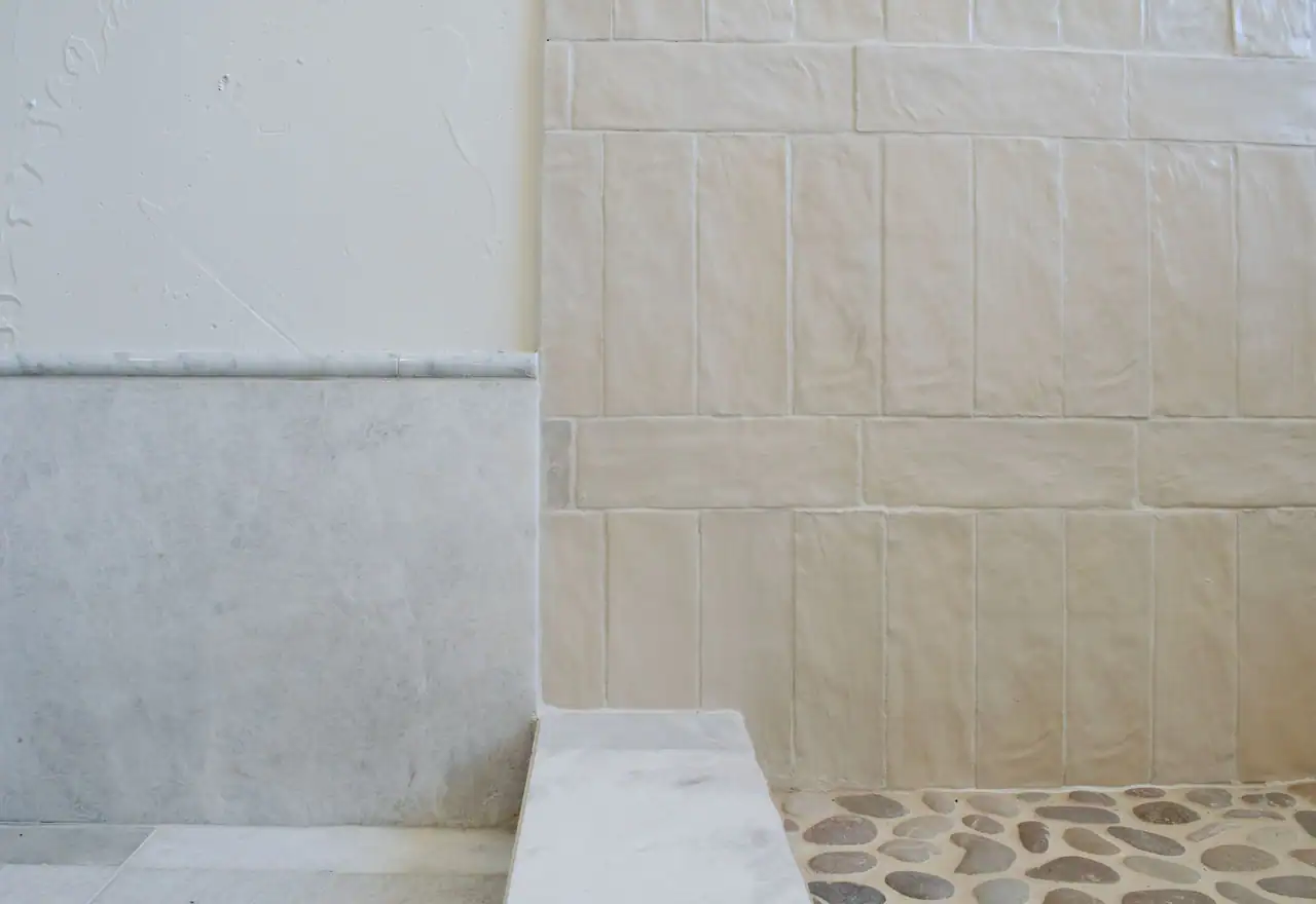 Plaster wall with gray-veined stone base meeting glazed white tile, a stone curb and a pebble shower floor