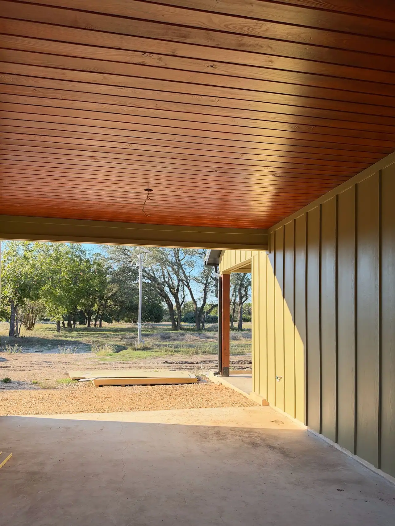 Covered porch with stained wood plank ceiling, olive board-and-batten wall and concrete floor, oak trees beyond