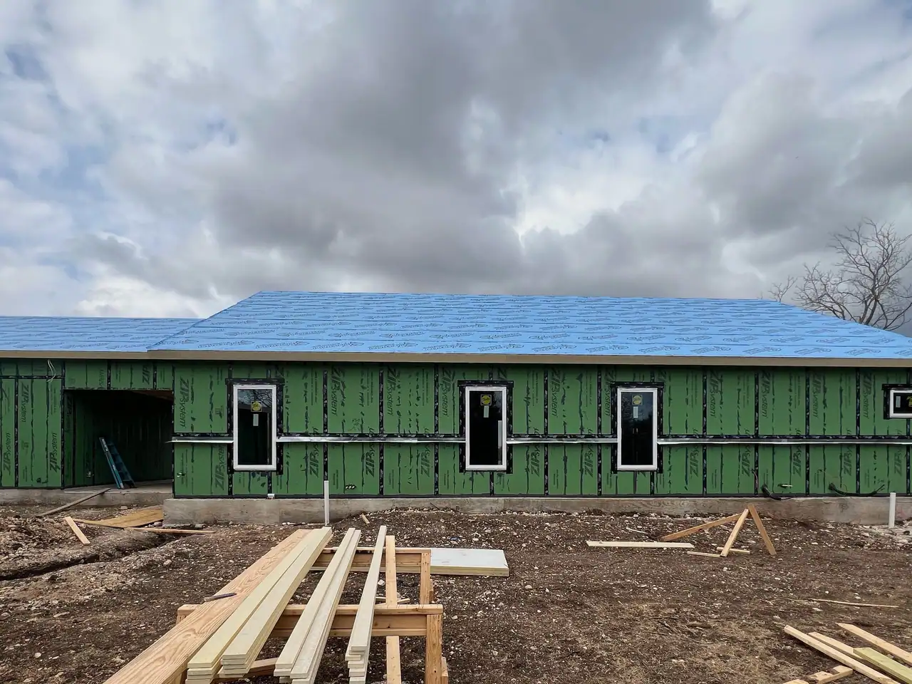 House in green ZIP sheathing with blue roof underlayment and installed windows, lumber on sawhorses in front
