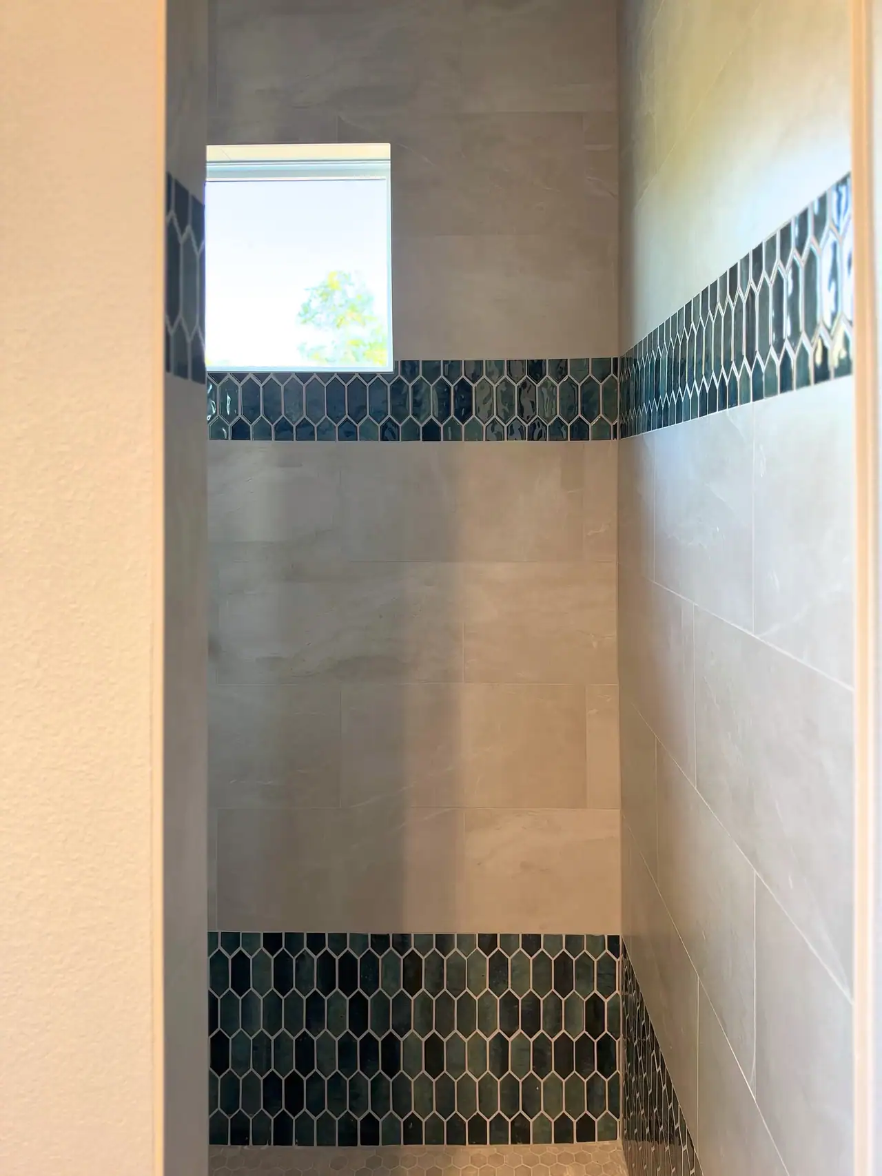 Walk-in shower with light gray tile, dark green elongated hexagon mosaic bands, hex floor tile and a window