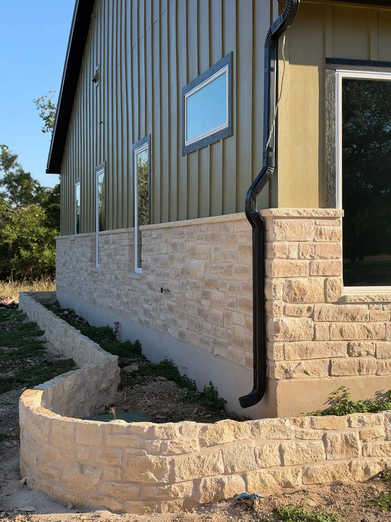 Curved low stone planter wall along a stone wainscot, olive board-and-batten siding, black downspout and windows