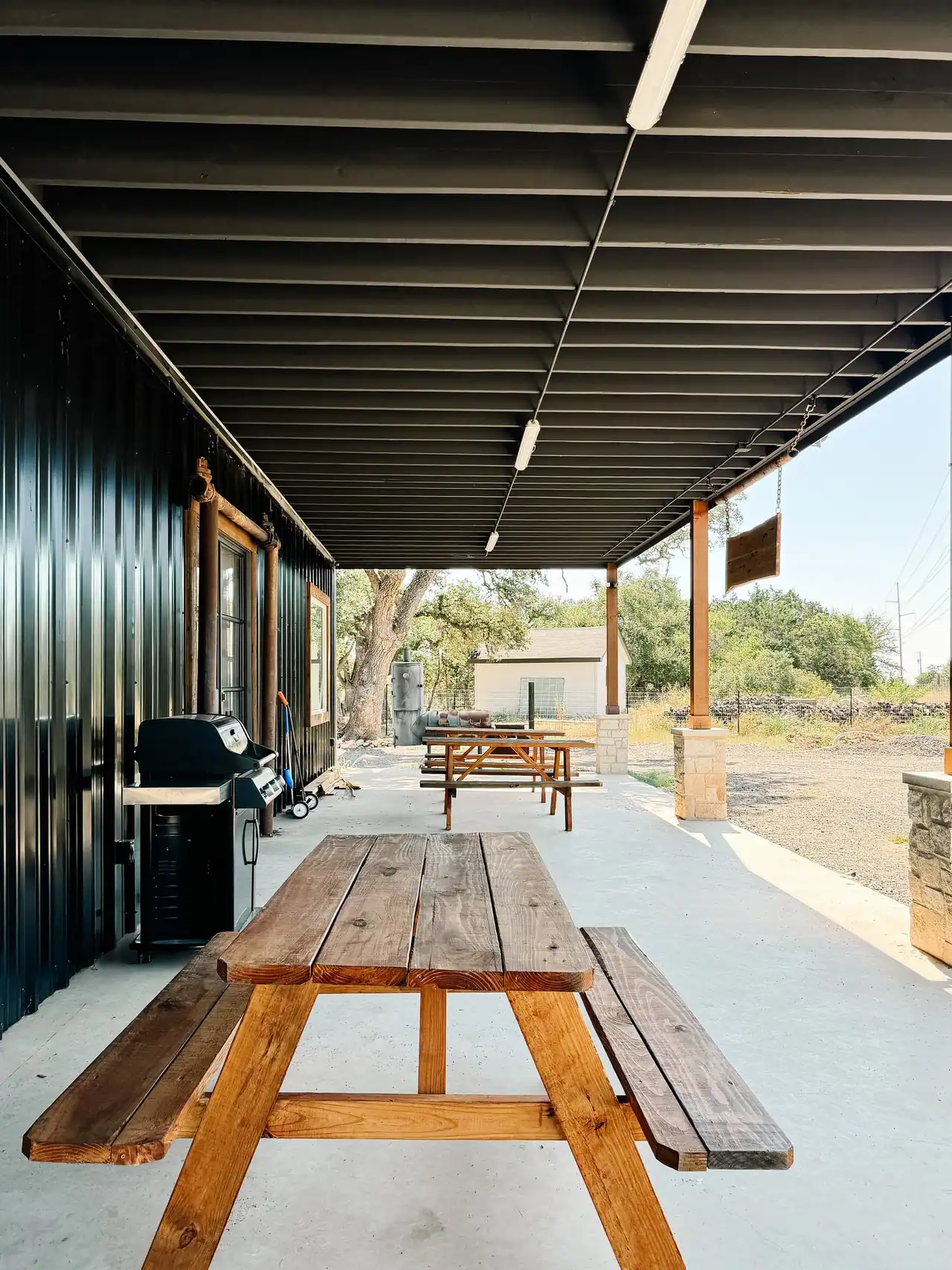 Covered patio with picnic tables and a grill beside a black metal wall, under dark joists, with timber posts on stone piers