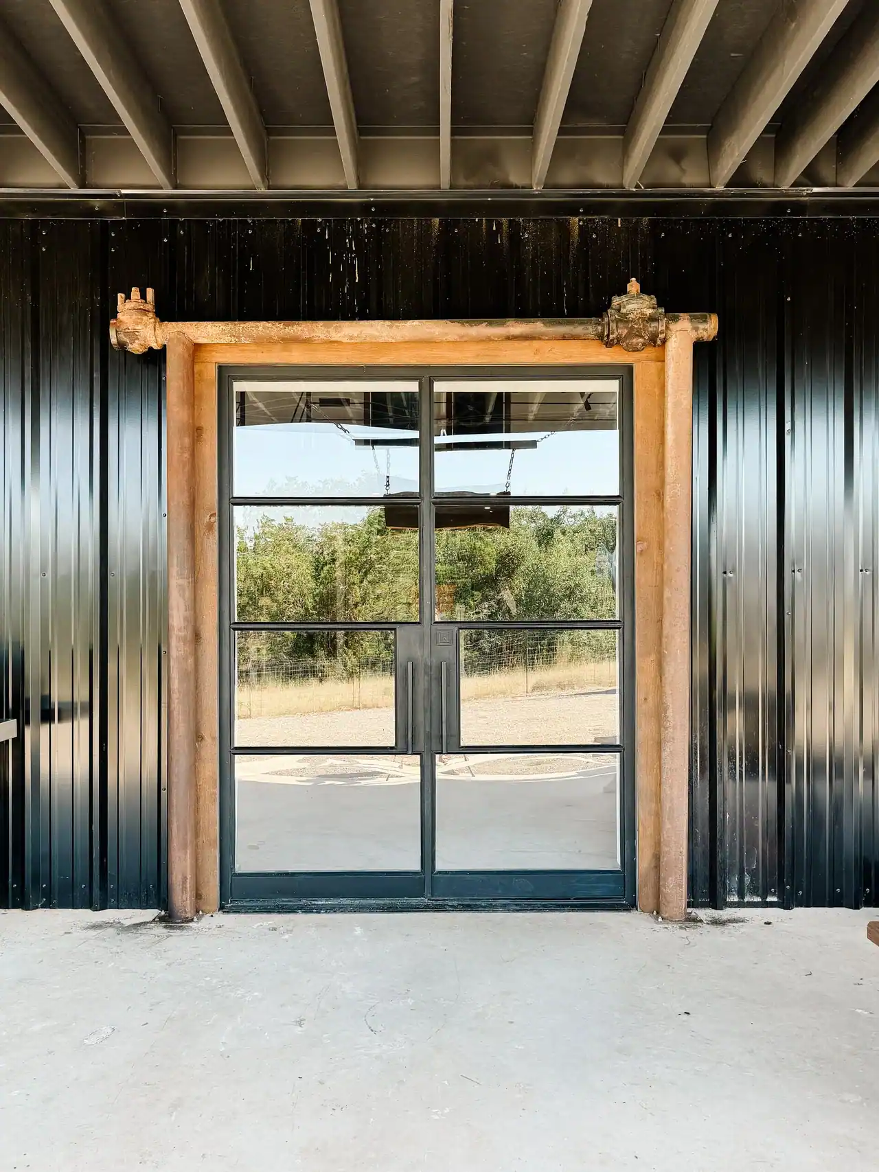 Black-framed glass double doors in a black metal wall, trimmed in rough timber with rusted pipe valves at the top corners