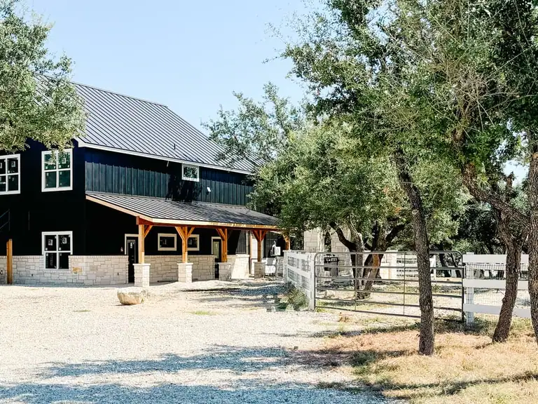 Black two-story barn with a timber porch on stone piers beside live oaks, a white fence and metal pipe gates