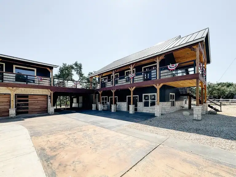 Black two-story barn and garage wing joined by an upper deck, with timber porches, stone bases and a stained concrete drive