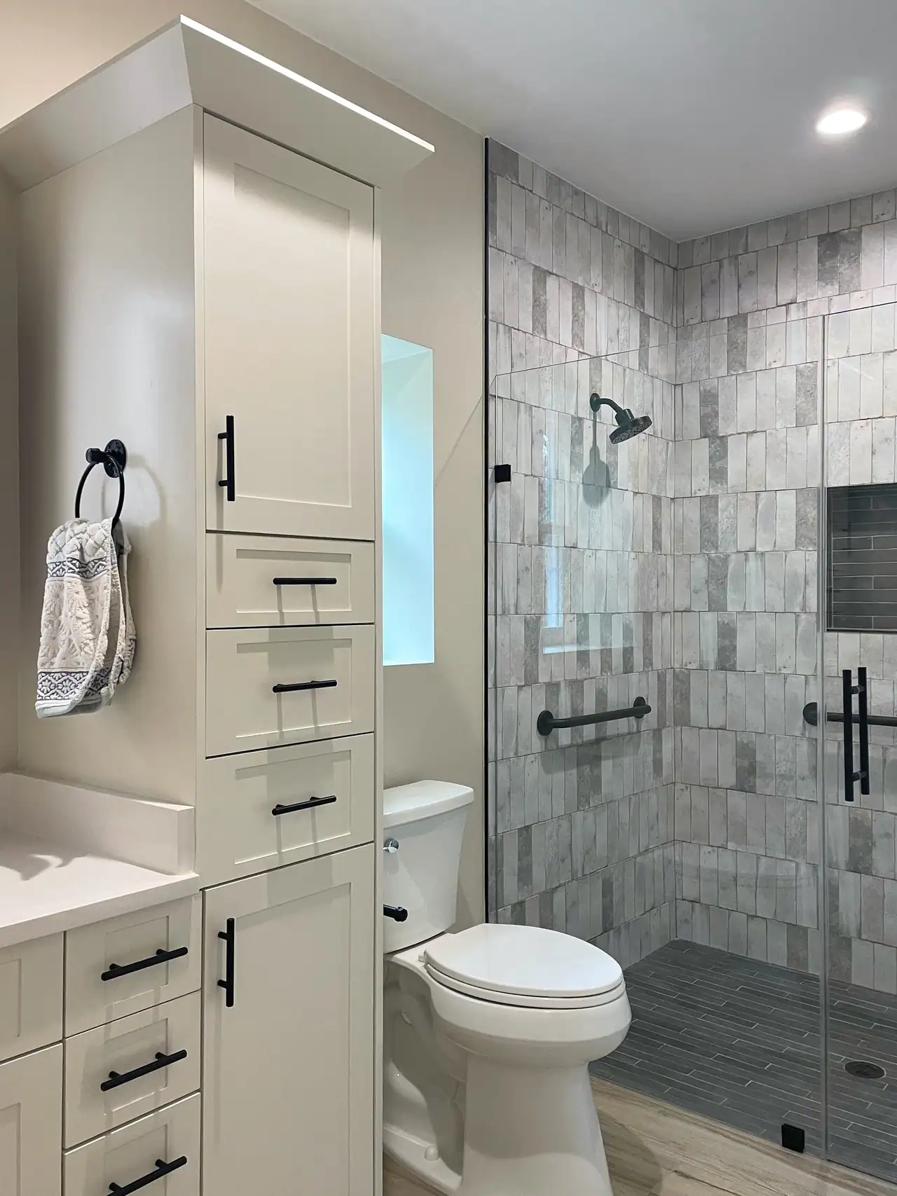 Bathroom with a tall white linen cabinet with black pulls, toilet and a glass walk-in shower in gray stacked tile