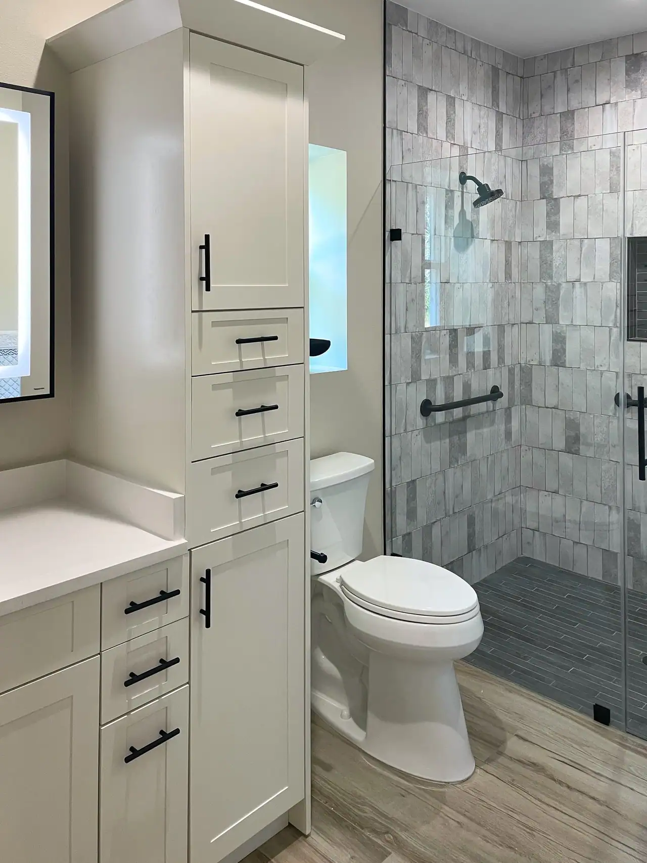 White linen tower with black pulls between a lit mirror and a toilet, with a glass walk-in shower in gray stacked tile