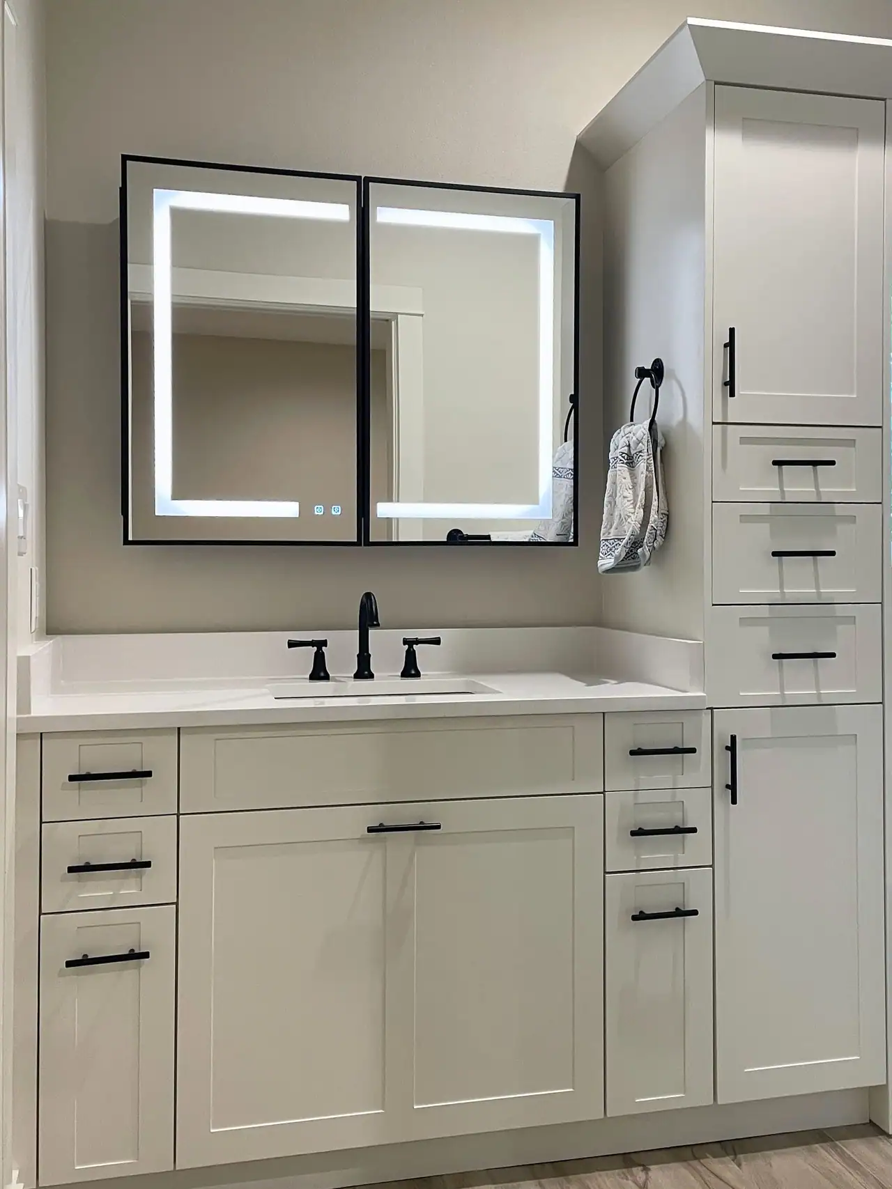 White shaker vanity with black faucet and pulls, a lit two-door mirror cabinet, tall linen tower and a window beyond