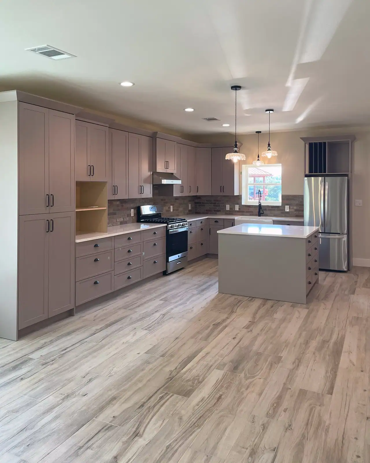 Open kitchen: taupe shaker cabinets, brick backsplash, gas range, island under glass pendants and a French-door fridge