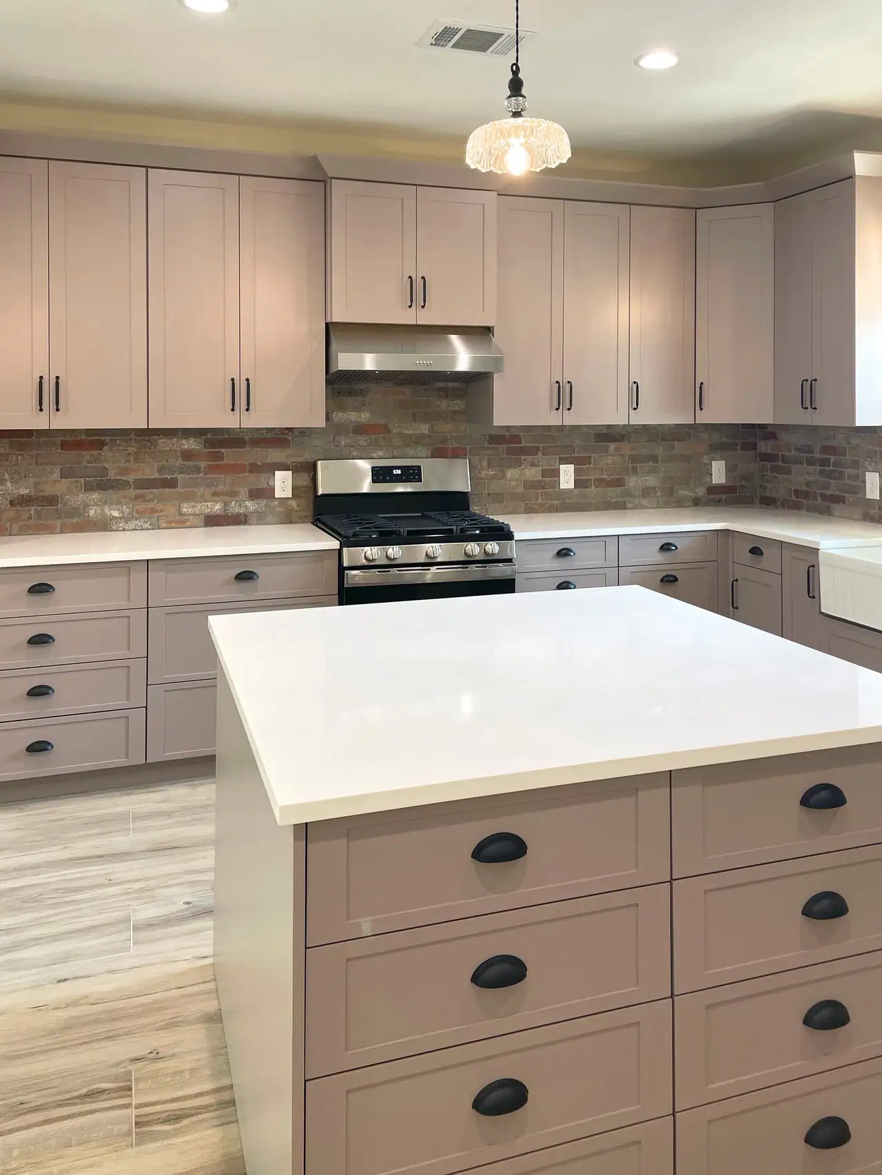 Kitchen island with a white top and taupe drawers with black cup pulls, gas range and brick backsplash behind, glass pendant