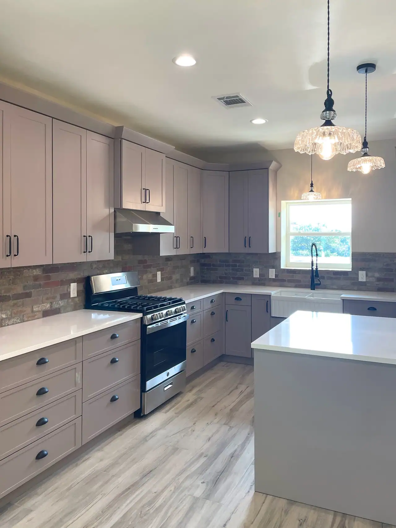 Kitchen with taupe shaker cabinets, brick backsplash, gas range and glass pendant lights over a white-topped island