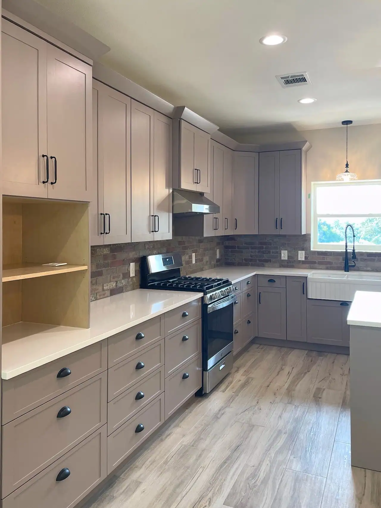 Kitchen with taupe shaker cabinets to the ceiling, brick backsplash, white counters, gas range and apron sink by a window