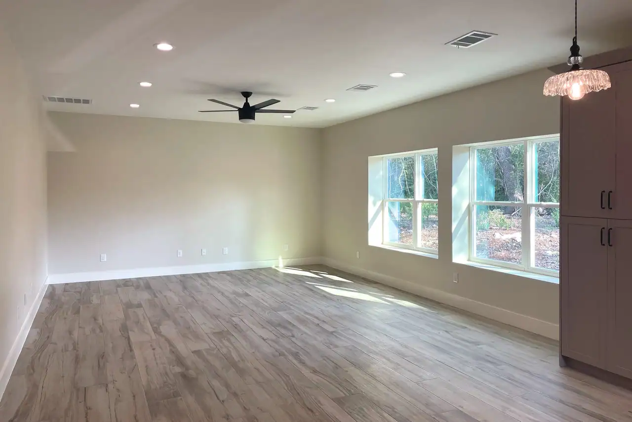Empty living room with wood-look plank floor, black ceiling fan, recessed lights and two large windows facing trees