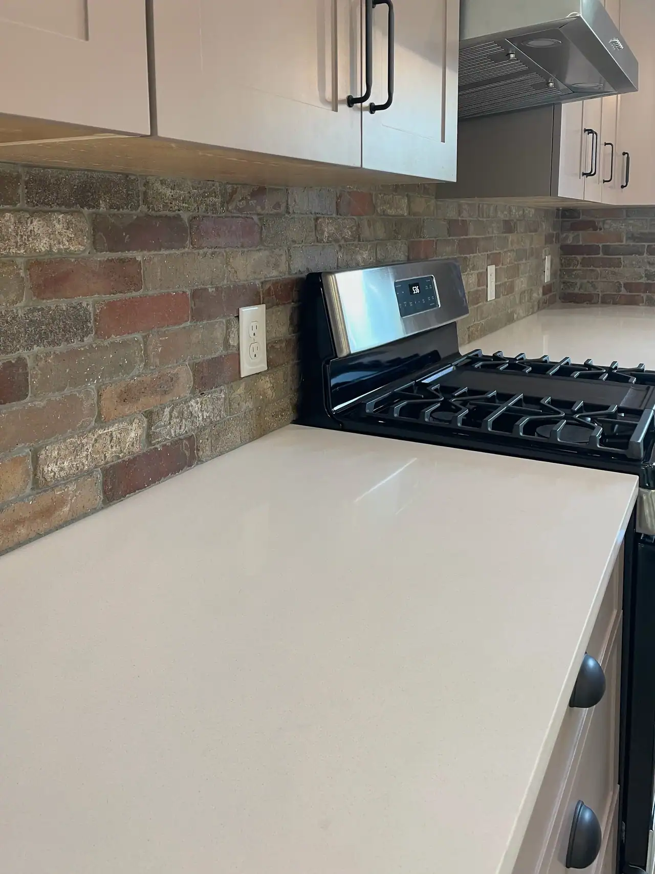 Close view of a brick backsplash, white countertop, gas range and taupe cabinets with black bar pulls and cup pulls