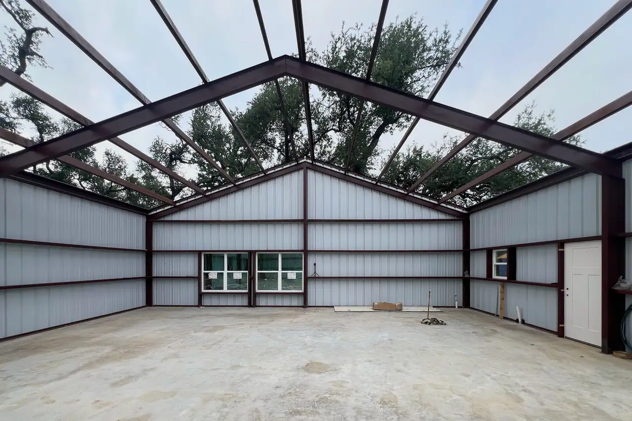 Dark red steel frame with light metal wall panels and open roof framing, oak branches overhead, a jobsite radio on the slab