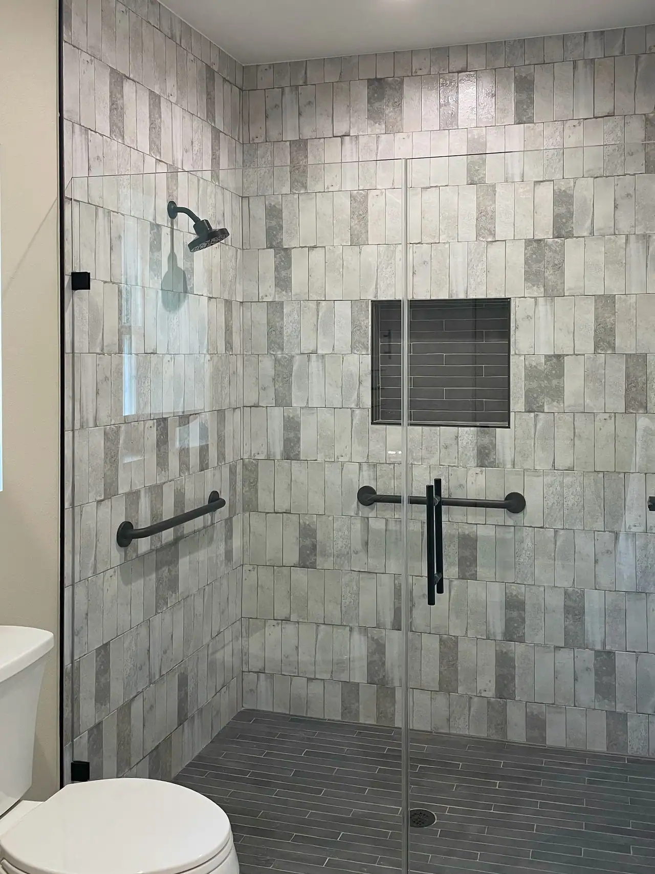 Walk-in shower behind glass with gray stacked tile, a recessed niche, dark floor tile, black shower head and grab bars