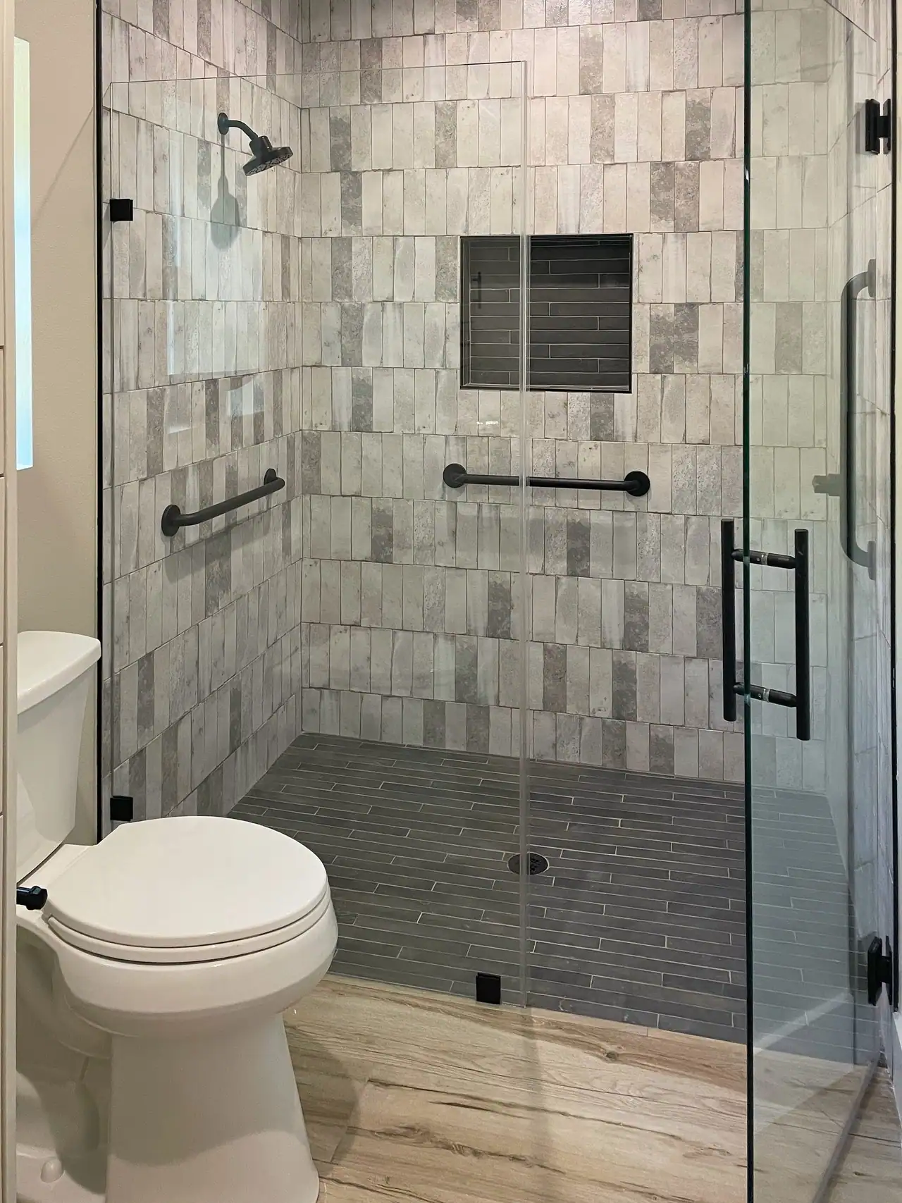 Glass walk-in shower with gray stacked tile, a dark tile niche and floor, black grab bars and door handle, toilet at left